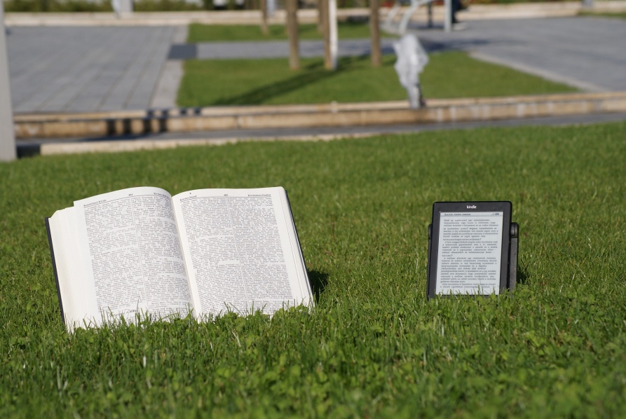 Tradycyjna książka z lewej, czytnik e-książek z prawej. Jedno i drugie obok siebie na trawie.