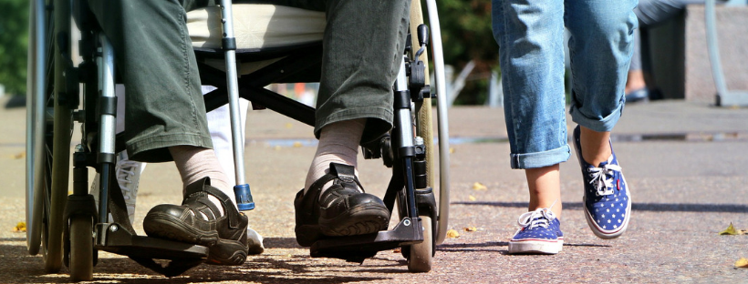dwie osoby na ulicy, jedna jest na wózku inwalidzkim, obok niej idzie druga osoba. Widzimy tylko ich nogi.