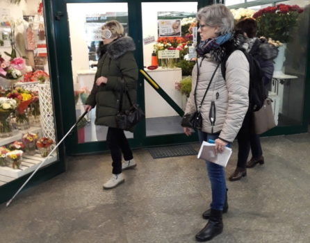 zdjęcie przedstawia dwie kobiety przed sklepem, jedna z nich idzie z laską i ma zasłonięte oczy, druga jej towarzyszy