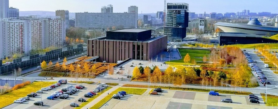 zdjęcie przedstawia część Katowic z loty ptaka. Można zobaczyć parkingi z samochodami, fragment stadionu Spodka, budynki biurowe. Pomiędzy nimi są jesienne piękne drzewa.