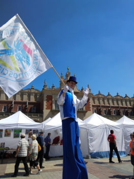 mężczyzna na szczudłach, ubrany w biało-niebieski strój z flagą Krakowa macha ręka. To chodząca reklama Kocham Kraków z wzajemnością.