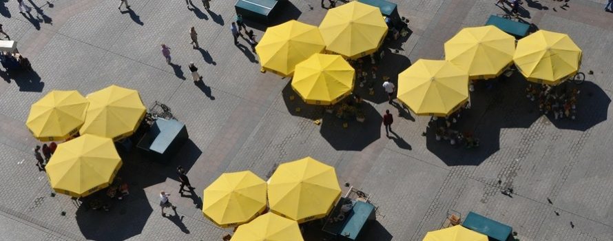 z lotu ptaka pokazane żółte duże parasole na rynku miasta. Pomiędzy nimi chodzą ludzie, poruszają się po gładkiej, równej nawierzchni.