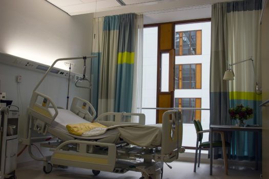 pokój szpitalny, po lewej stronie stoi łóżko szpitalne, w prawym rogu krzesło przy stoliku, na stoilku stoi bukiet kwiatów; w tle znajduje się okno; światło przygaszone, panuje półmrok