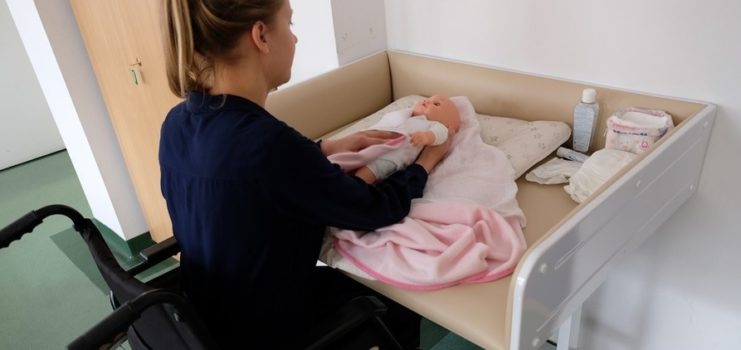 młoda kobieta na wózku siedzi przy przewijaku dla niemowląt. Leży na nim lalka, a kobieta uczy się ja przewijać. Kobiety z niepełnosprawnościami ze szkoły rodzenia Bocianie gniazdo w szpitalu na Solcu w Warszawie mogą się uczyć.