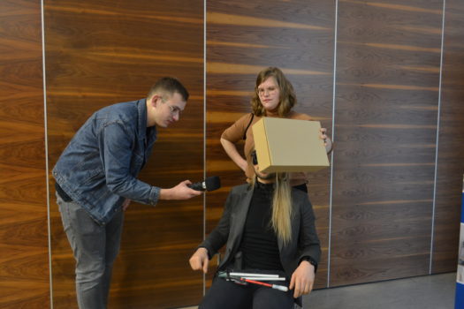 instruktorka trzyma tekturowe pudło przed siedzącą na krześle osobą. Obok stoi dziennikarz radia Eska i nagrywa