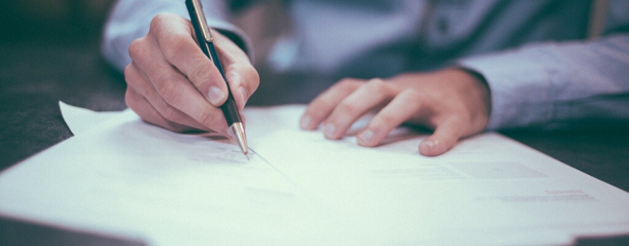 fragment męskiej sylwetki. mężczyzna ubrany w niebieską koszulę w prawej dłoni trzyma pióro do pisania, przed nim kilka białych kartek, które podpisuje