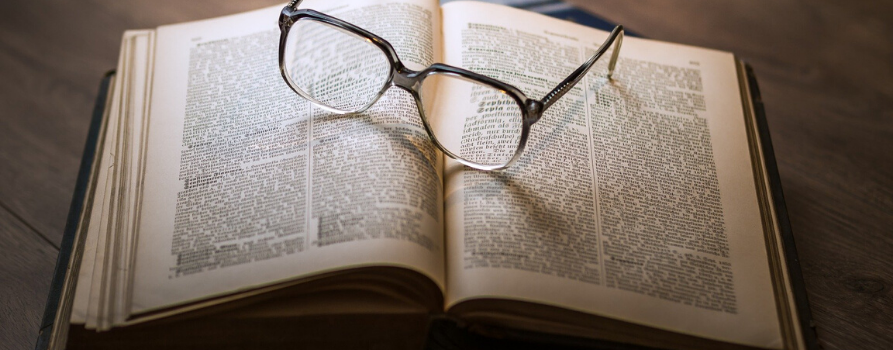 na drewnianym stole leży otwarta książka. na książce leżą okulary.