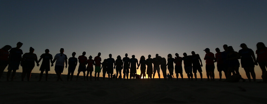 zachód słońca, które jest już prawie przy samym horyzoncie. na tle tego nieba widać kontury około 20 osobowej grupy ludzi, która trzyma się za ręce.