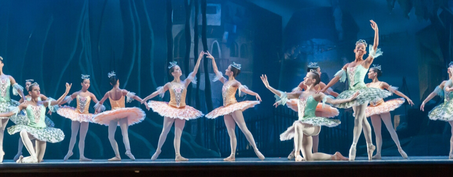 scena teatralna. na tle niebieskiej scenografii tańczy kilkanaście baletnic ubranych w pastelowe stroje.