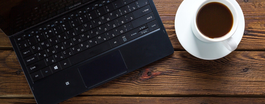 fragment klawiatury czarnego laptopa - obok po prawej biała filiżanka z kawą