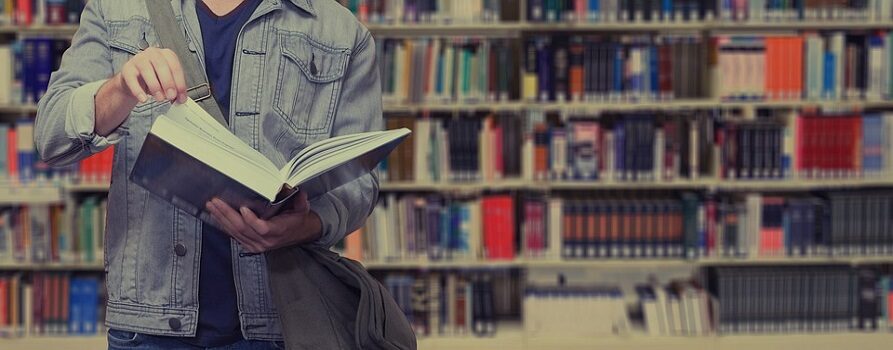 Student trzymający w rękach otworzoną książkę, stojący na tle regałów z książkami, prawdopodobnie w bibliotece
