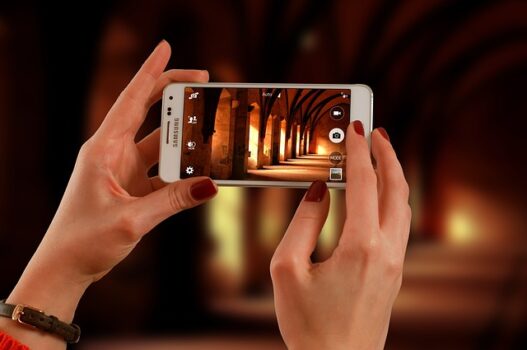 Kobiece dłonie, trzymające smartfon. Na ekranie telefonu zdjęcie łukowatych sklepień oraz filarów. Prawdopodobnie wnętrze kościoła.