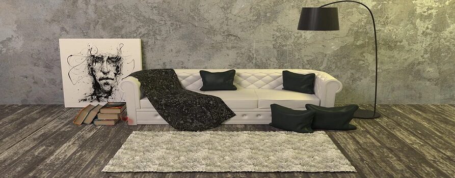 Salon, pośrodku szara kanapa, na niej dwie czarne poduszki i czarny koc, po prawej stronie czarna lampa, przed sofą dywan, po lewej stronie obraz oparty na ścianie