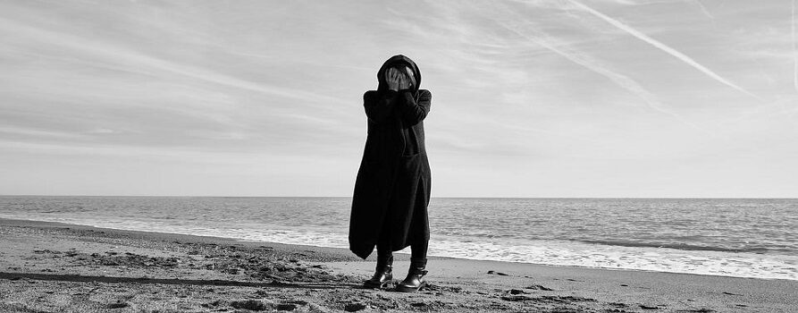 Czarno białe zdjęcie kobiety, ubranej w długi dres z kapturem, stojącej na plaży. Kobieta zakrywa twarz rękoma geście rozpaczy