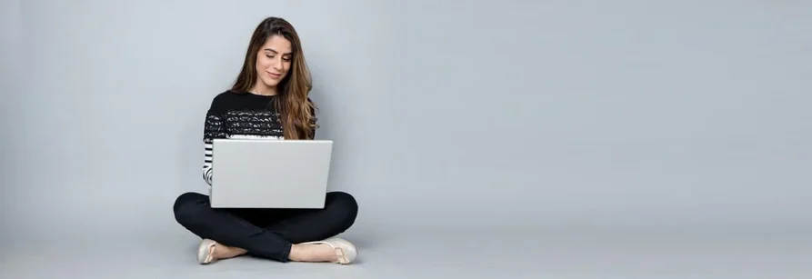 Młoda dziewczyna siedząca na podłodze w siadzie skrzyżnym, pracująca na laptopie.