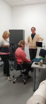 Delegacja z Danii w Ośrodku Wsparcia i Testów w Laskach. Dwie kobiety, jedna siedzi na krześle obrotowym przed komputerem, druga stoi za nią. Przed nimi mężczyzna, opowiadający o Ośrodku